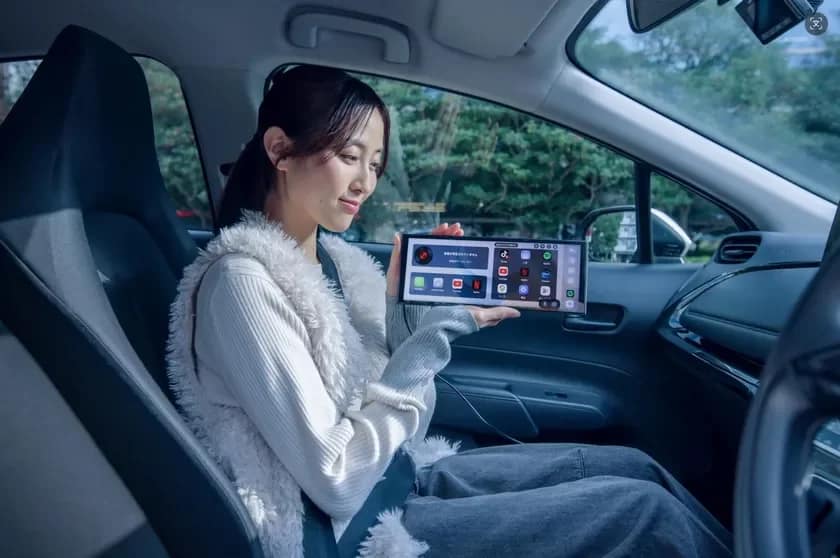 車の助手席に座る若い女性が、様々なアプリが表示された大型のポータブルスクリーンデバイスを手に持っています