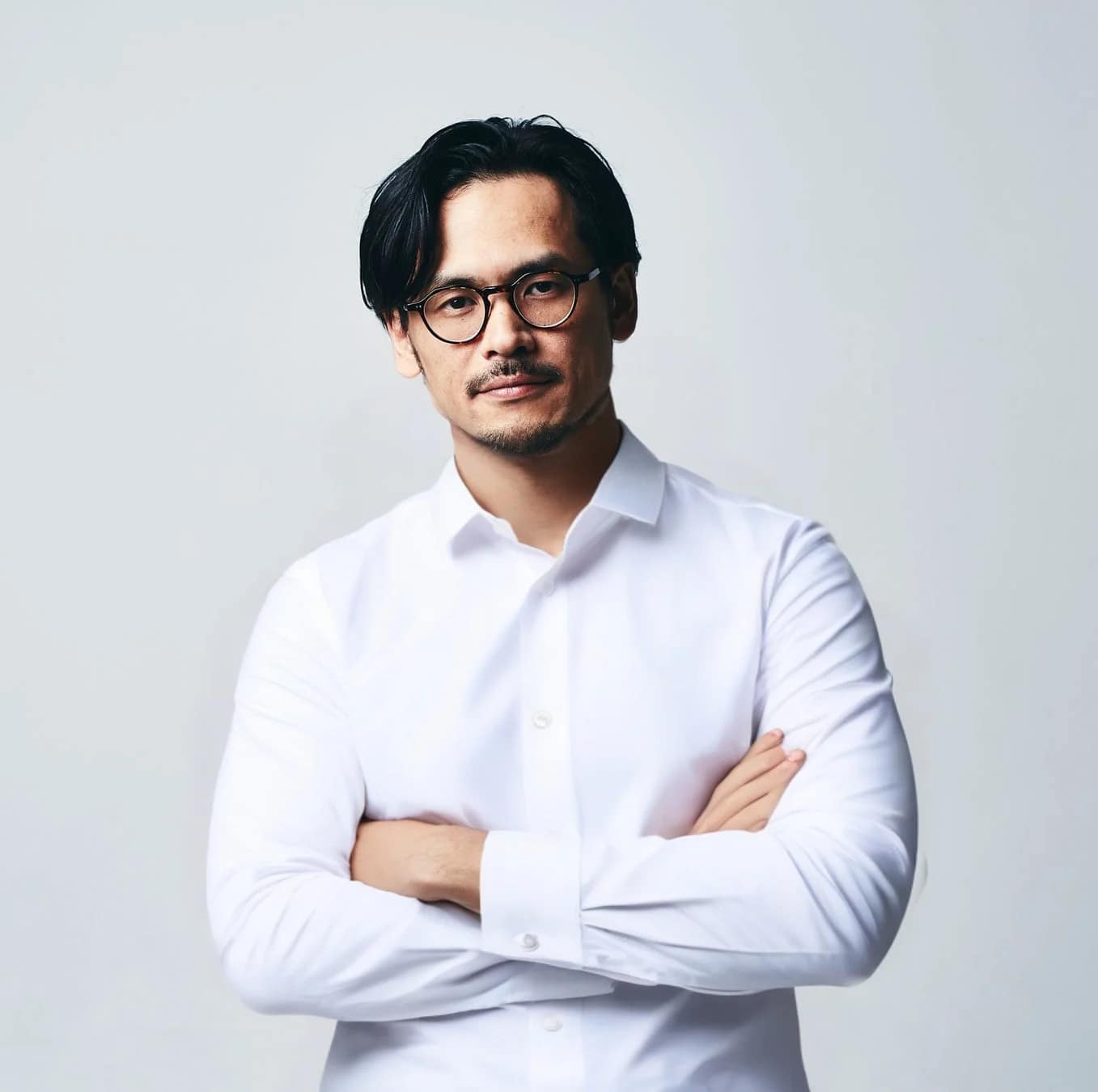 眼鏡をかけたアジア人男性が白いシャツを着て腕組みをし、真剣な表情でカメラを見つめている上半身のポートレート写真。背景はシンプルな白です。