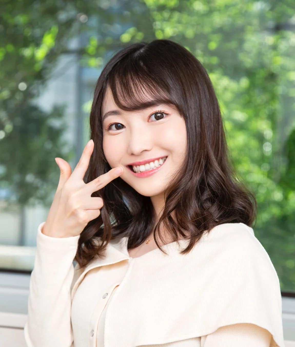満面の笑みを浮かべた若い女性が、ピースサインのように指で口元を指しているポートレート。背景には緑の葉がぼやけて写っており、明るく爽やかな印象を与えます。
