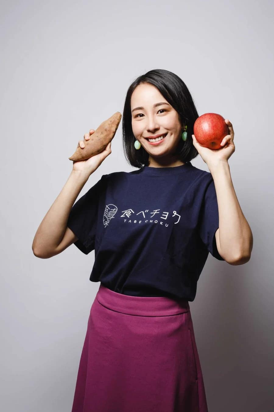 笑顔の女性がサツマイモとリンゴを両手に持ち、「食べチョク」のロゴ入りTシャツを着て立っているスタジオ写真です。