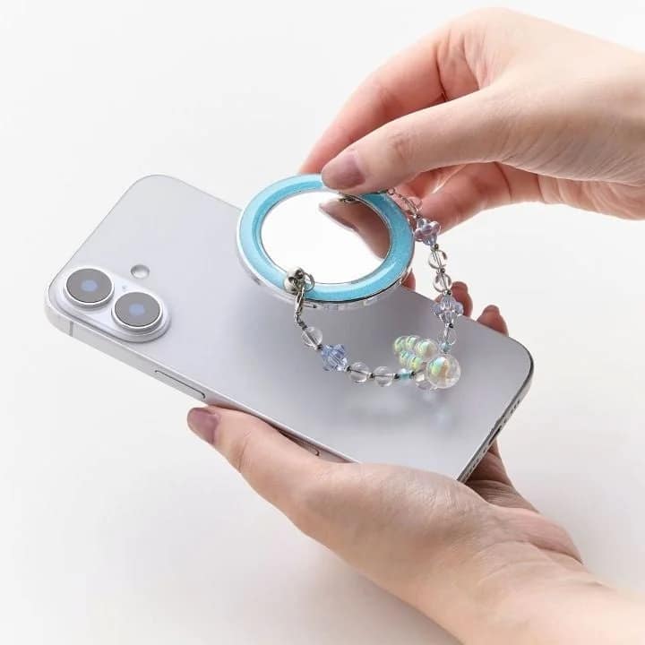 人物がシルバーのスマートフォンを手に持っており、背面には水色の縁取りとビーズのストラップが付いた丸いミラー型アクセサリーが装着されている様子