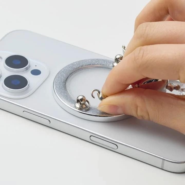 グリッター加工されたシルバーのリング型アタッチメントが付いたスマートフォンの背面に、手が小さなシルバーのチャームを取り付けている様子