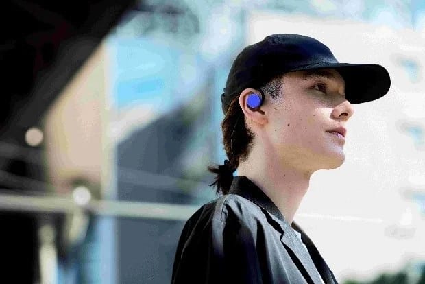 黒いキャップをかぶり、青いワイヤレスイヤホンを装着した若い男性が、屋外で空を見上げるようなポーズをとっています。
